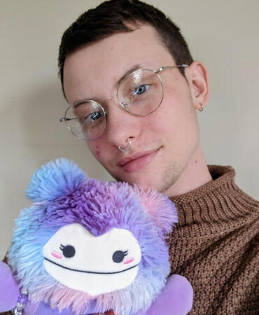 Photo of me, a white enby with short brown hair, glasses, septum piercing, and a purple bigfoot squishmallow named Princess Autism!